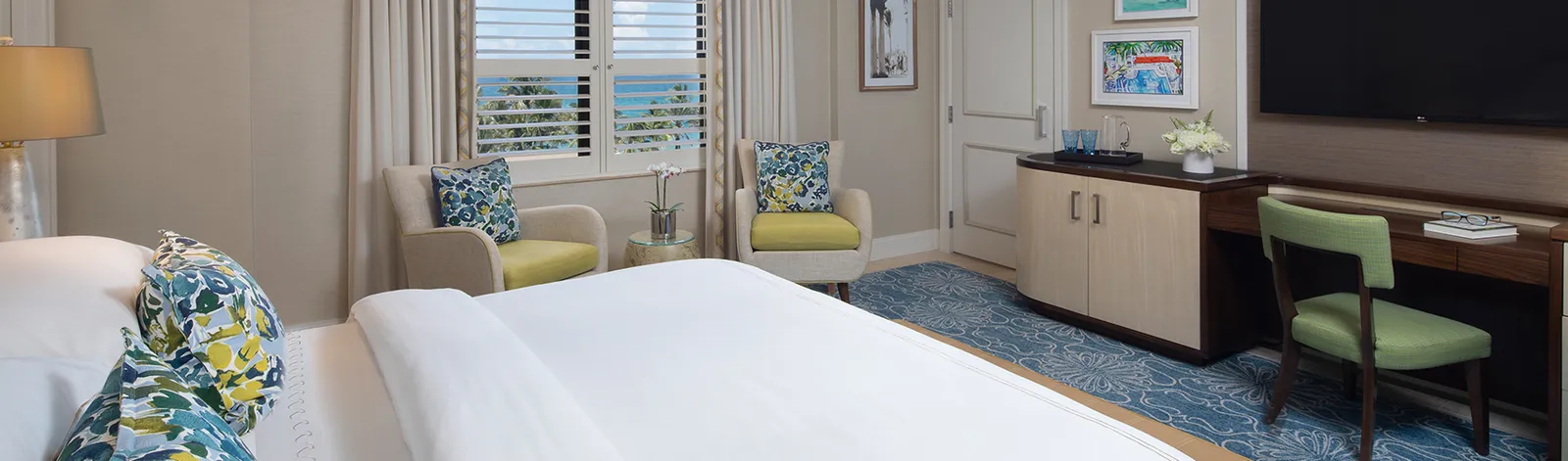 A Deluxe Guest Room: the bed in front, 2 small armchairs under the shuttered window and the desk with the television.