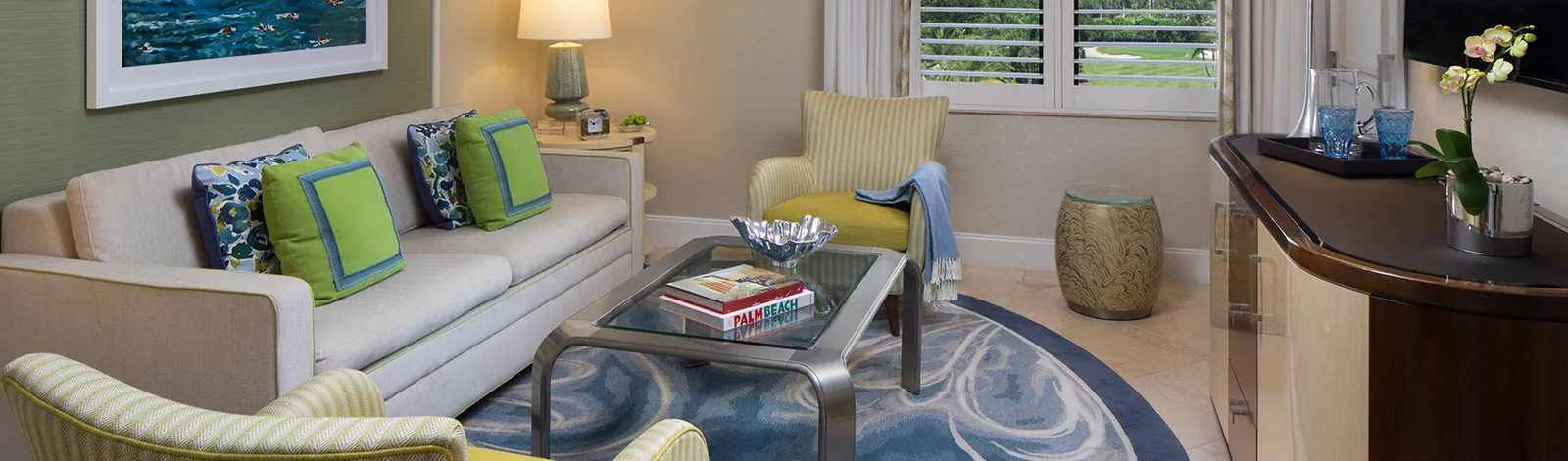 A Deluxe Suite’s sitting room: the sofa with green and blue cushions on a blue rug, a yellow armchair and the white-shuttered window.