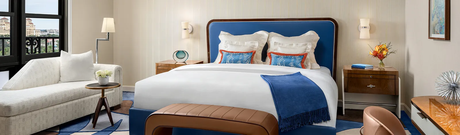 A Flagler Club Guest Room: the bed with its blue headboard and leather bench, a white chaise longue and the window on the resort side.