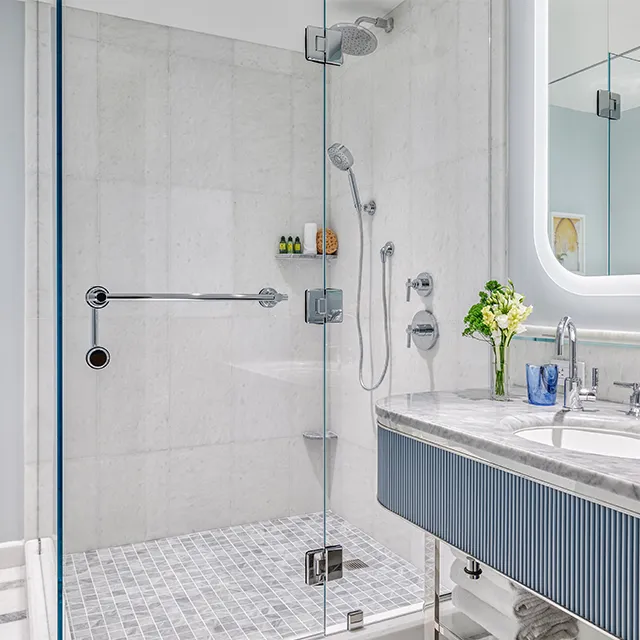 A Flagler Club Guest Room’s bathroom: the pale marble shower with its rainfall head behind glass, and the basin under a lit mirror.