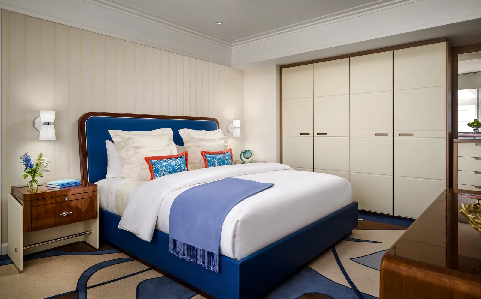 A Flagler Club Signature Suite’s bedroom: the bed with its blue headboard, coral cushions and a wall of cream wardrobes.
