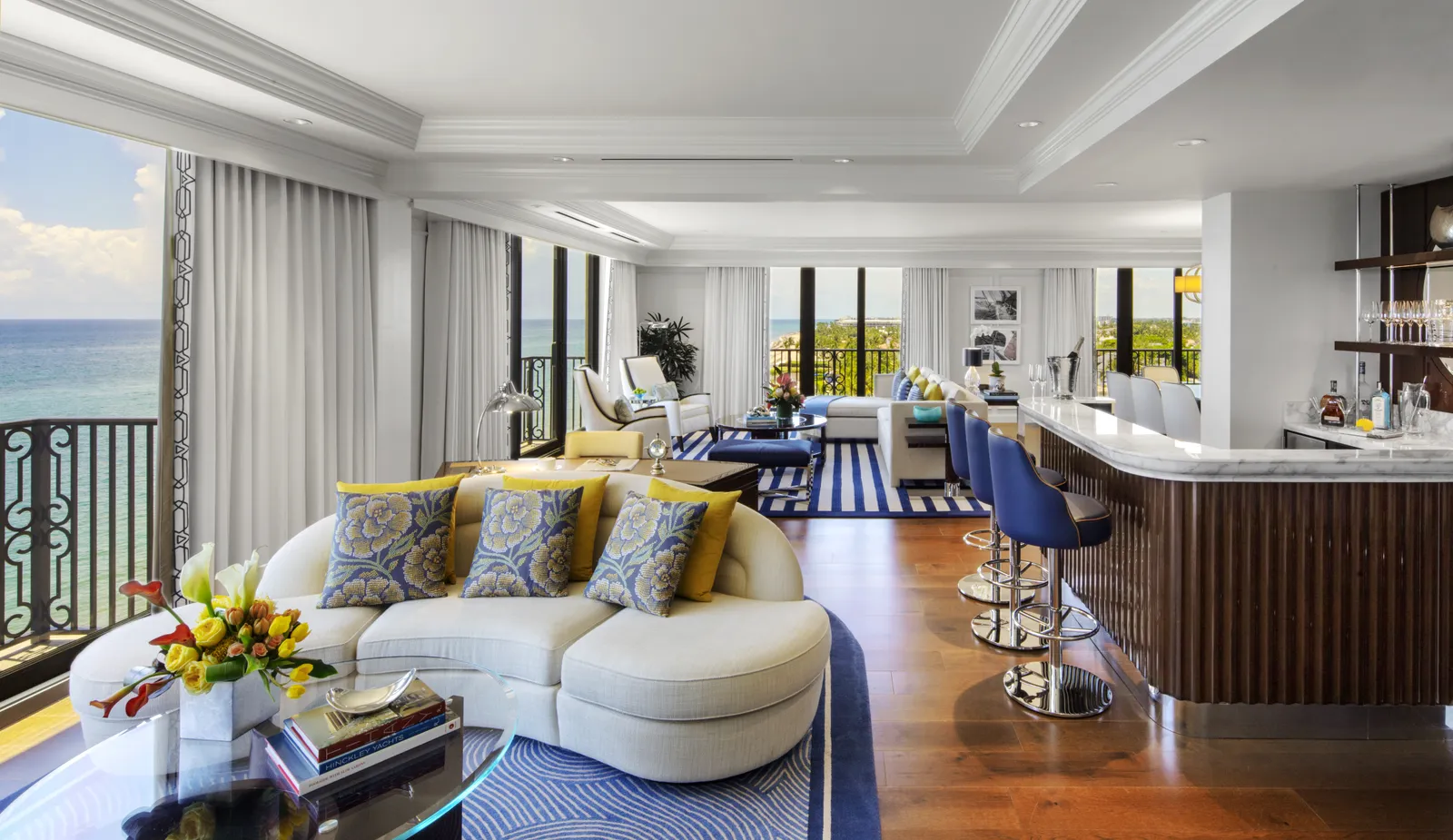 The Imperial Suite’s living room: a curved cream sofa, the bar counter with blue stools and glass doors onto the ocean on 2 sides.