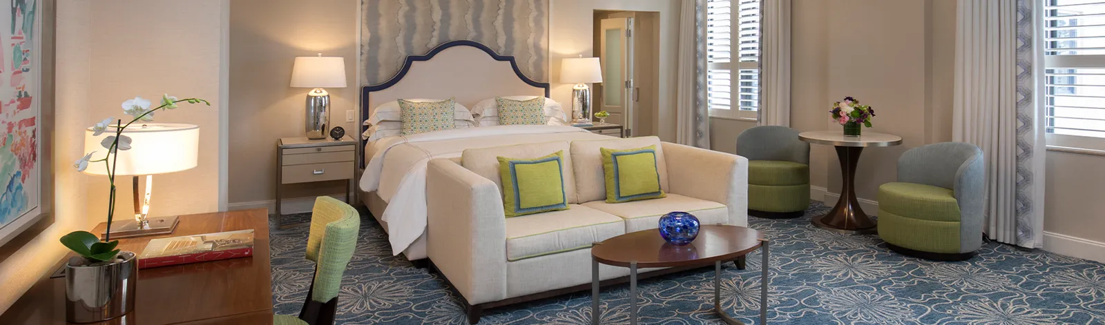 A Junior Suite: the king bed, a small sofa at its foot, 2 grey armchairs round a table and shutters at the window.