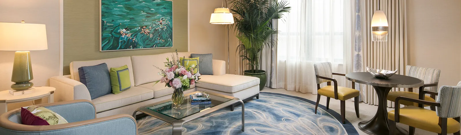 The living room of a Suite with Limited View: the corner sofa, a round table with yellow chairs and pale curtains at the windows.