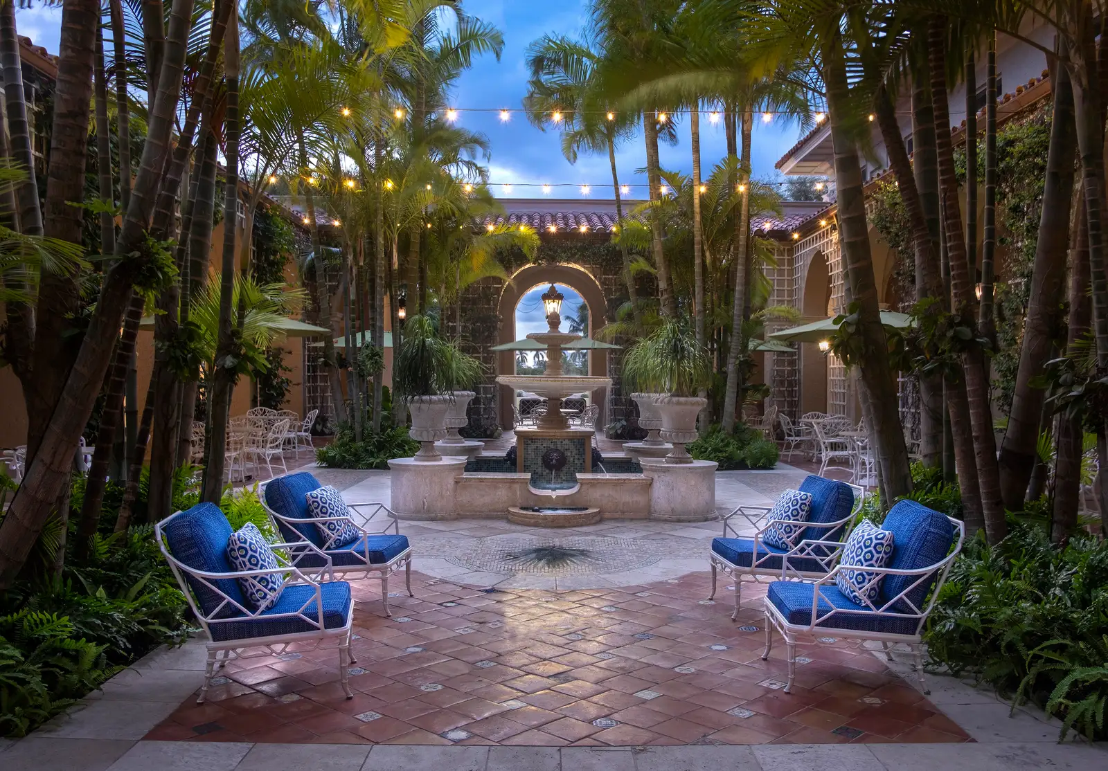 The Palm Courtyard at dusk: a stone fountain in the middle, white iron armchairs with blue cushions, palms and a string of lights.