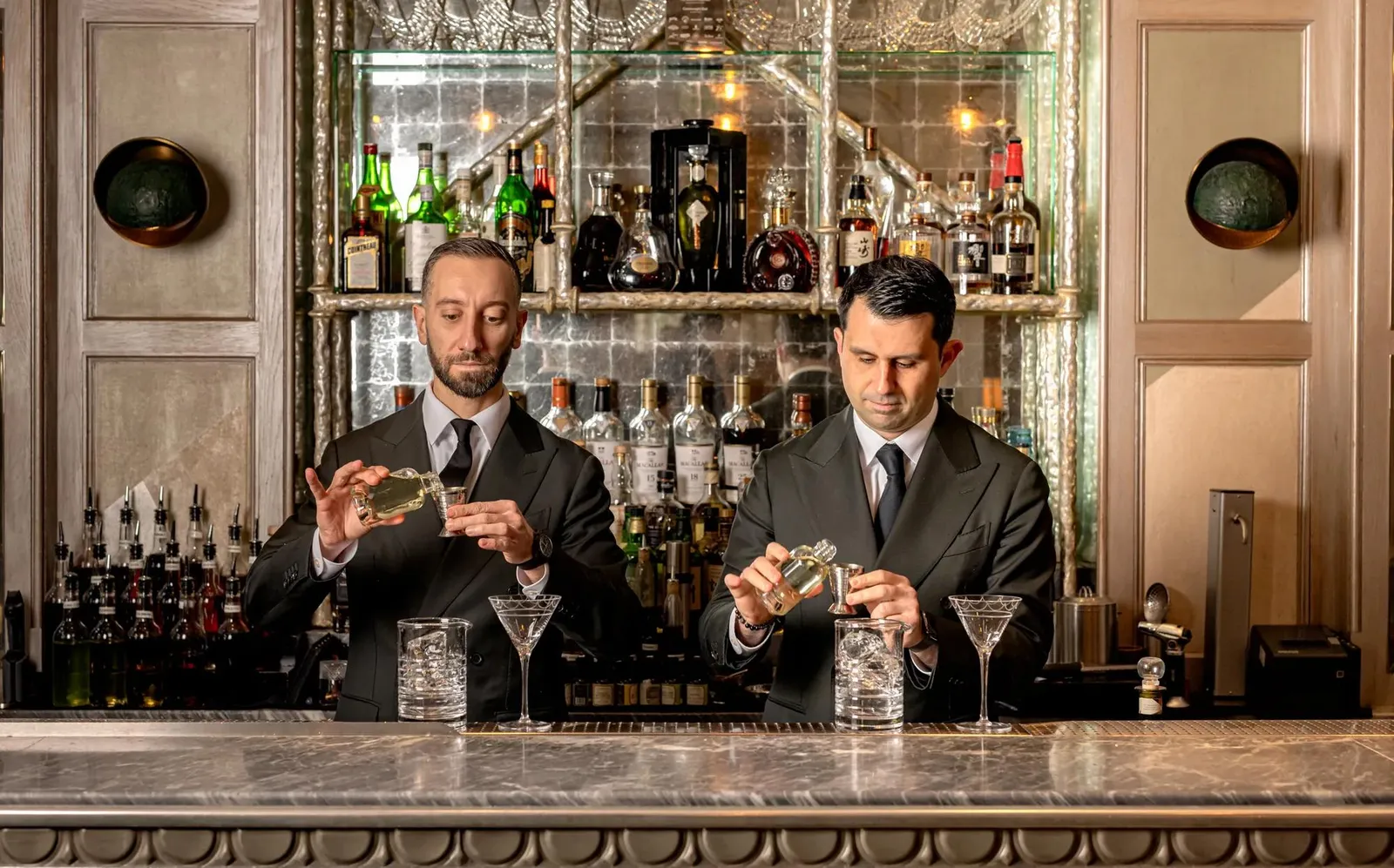 2 bartenders in jackets make Martinis at the Connaught Bar counter, among bottles and mirrors