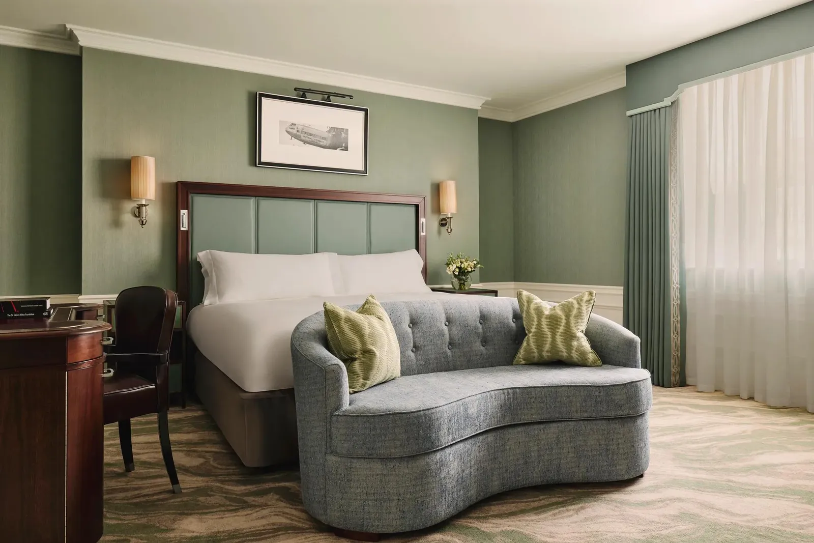 A Courtyard Deluxe Room: a bed with a duck-egg headboard, a grey loveseat and pale curtains