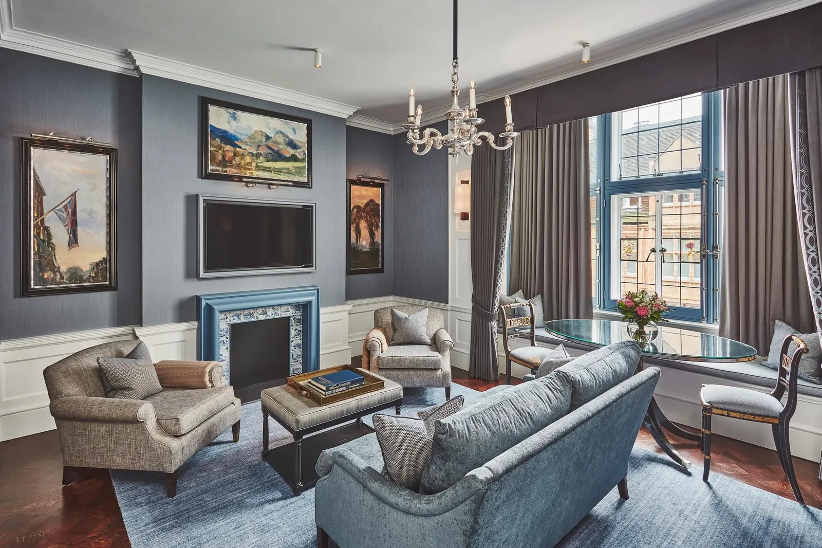 The Two-Bedroom Grosvenor Suite: a blue sitting room with a tiled fireplace, a chandelier and a dining table by the window