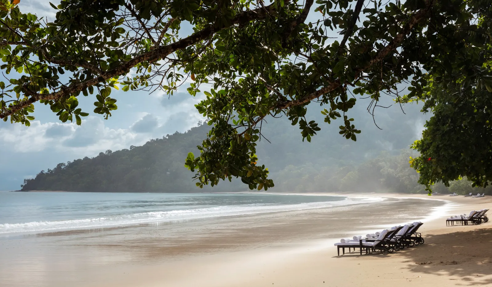 2 loungers under the trees at the end of Datai Bay’s beach, the headland’s hills across the water
