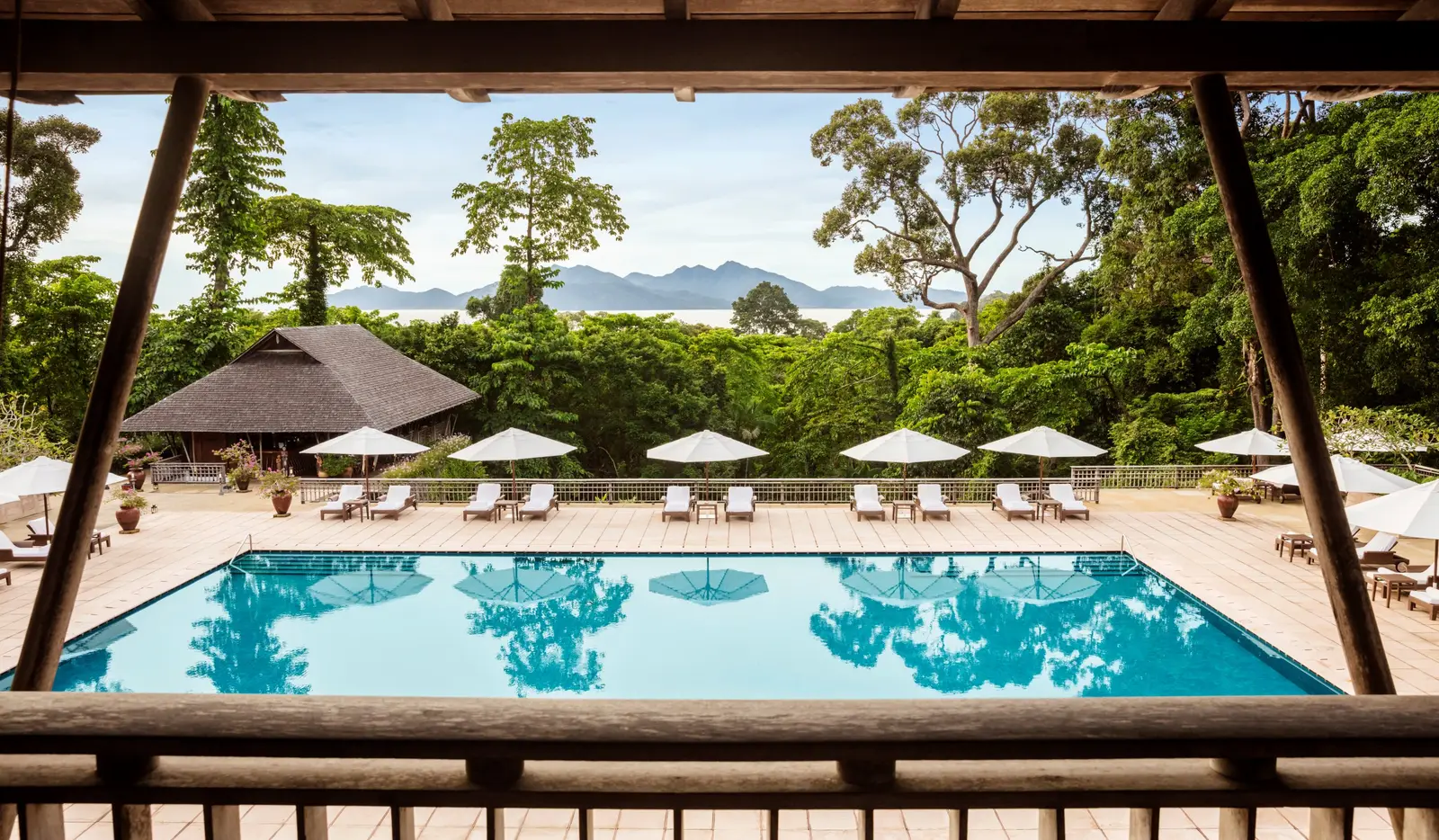 The main pool seen between the lobby’s timber posts, umbrellas along its edge, forest and bay behind