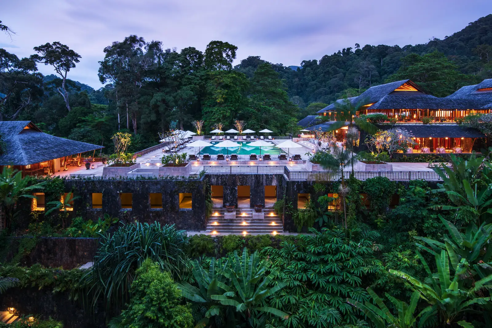 The main building and the lit pool at dusk, seen from above the forest