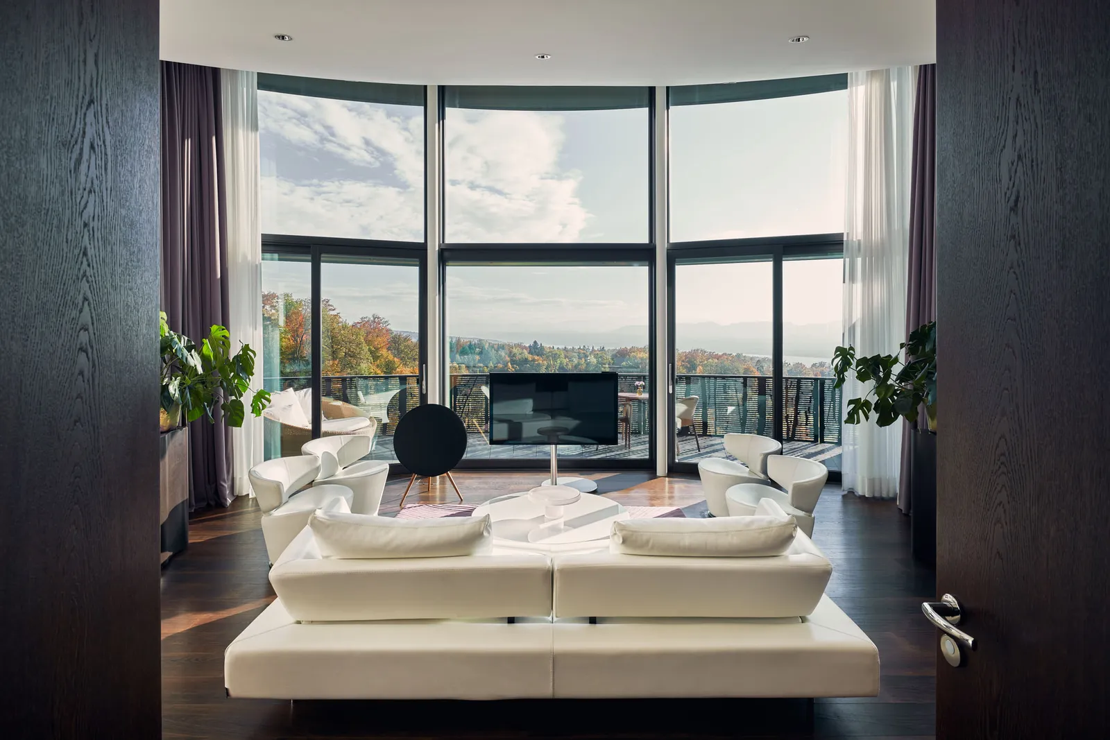 The living room of the Golf Suite: white sofas facing a curved wall of glass onto the terrace and the autumn trees