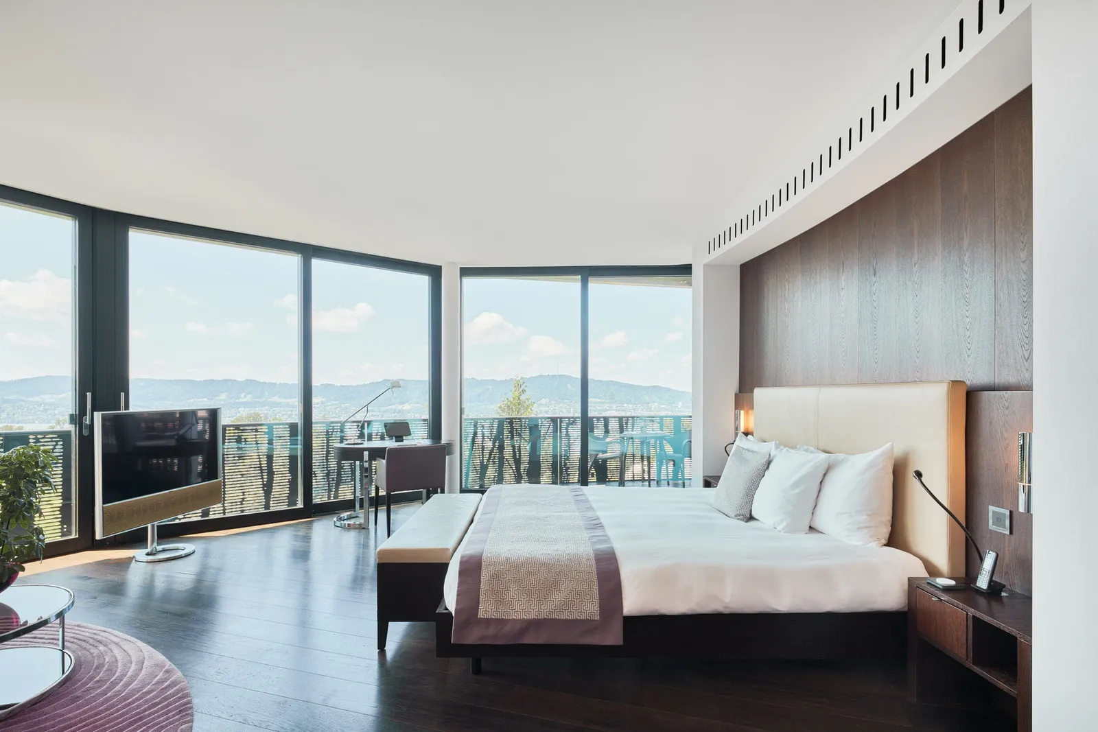 A Junior Suite Grand in the wing: the bed and a curved wall of windows onto the balcony and the hills