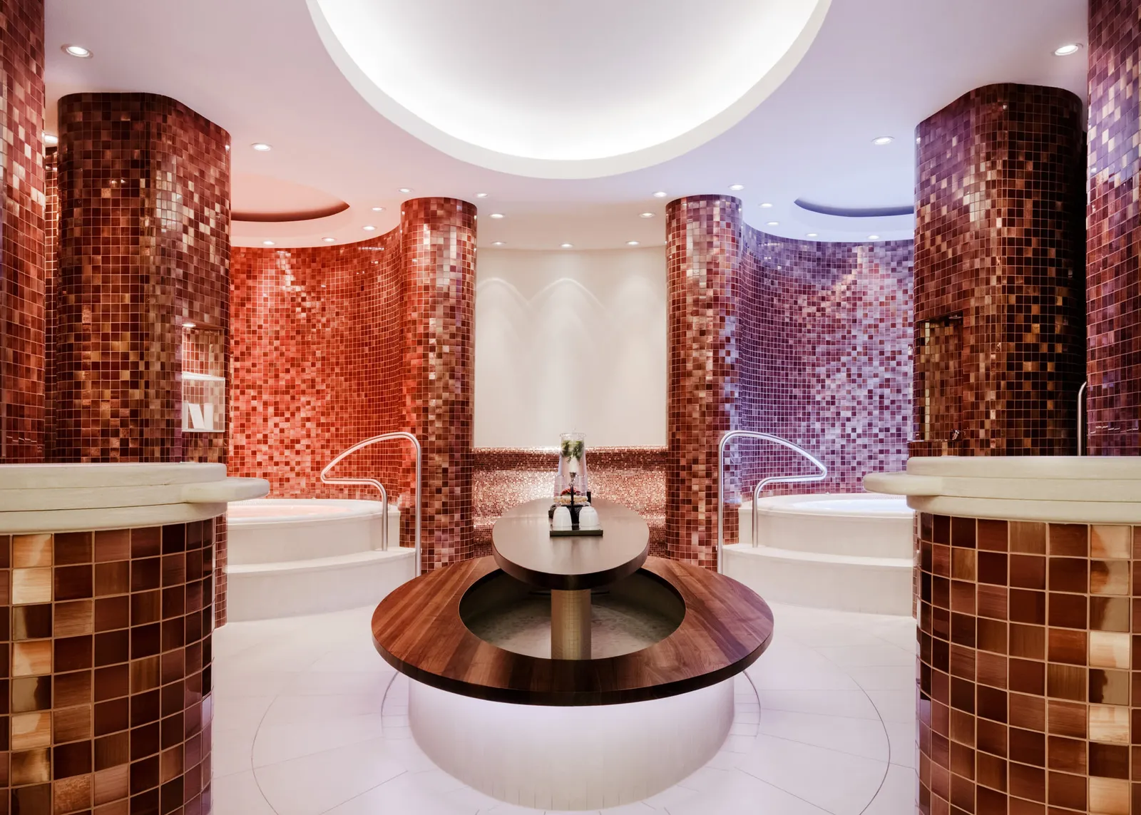 A round room in the spa clad in red and violet mosaic, with basins around a wooden central fountain