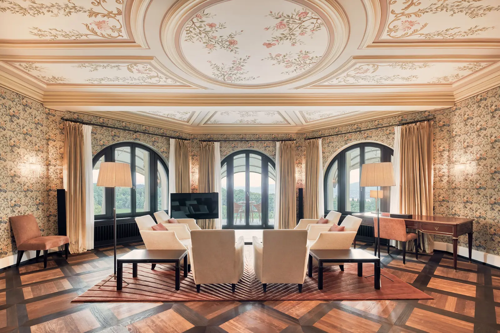 The living room of a Suite Deluxe: a painted stucco ceiling, floral wallpaper and 3 arched windows