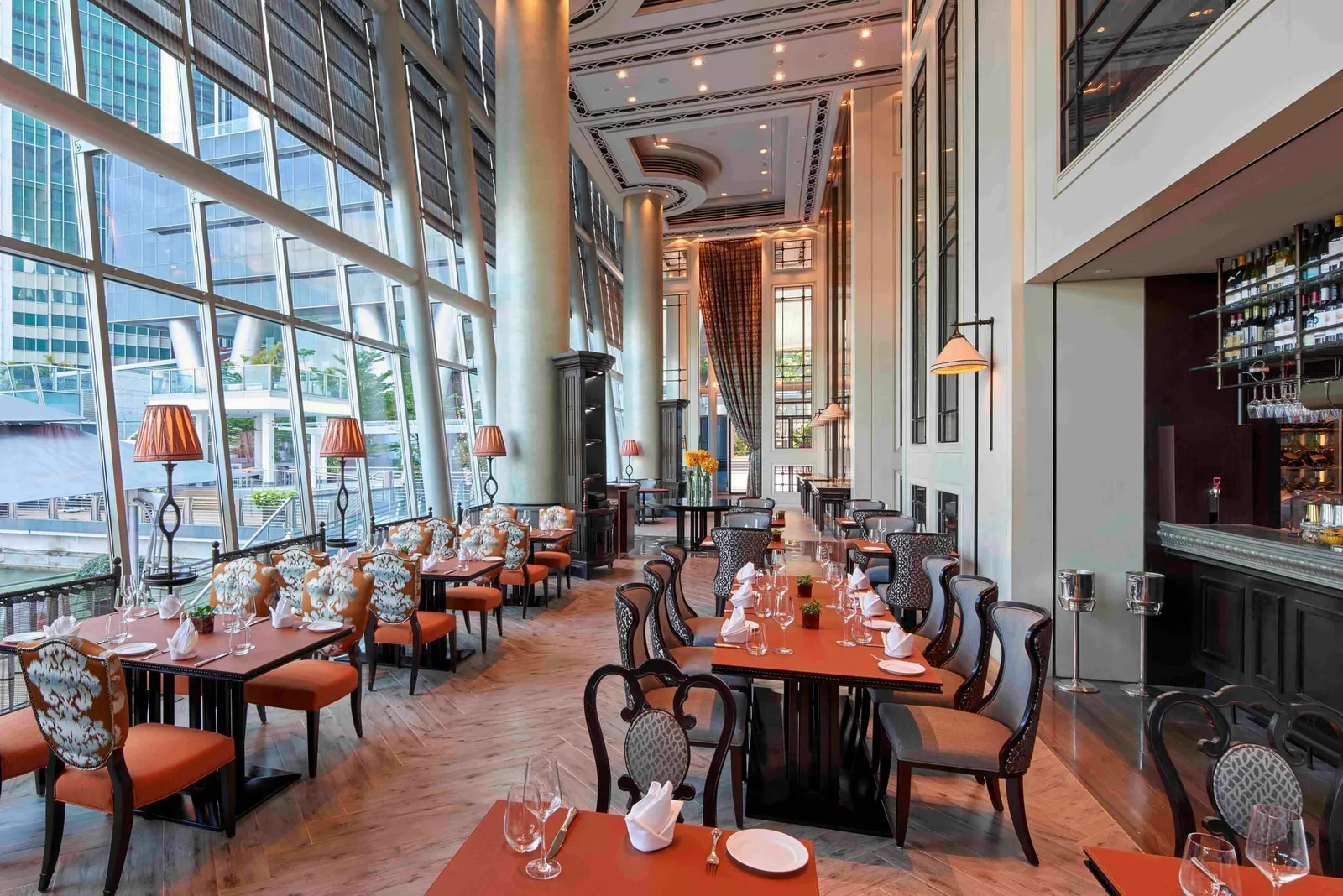 La Brasserie by day: laid tables along a wall of windows 2 storeys high, orange-backed chairs and the bar counter.