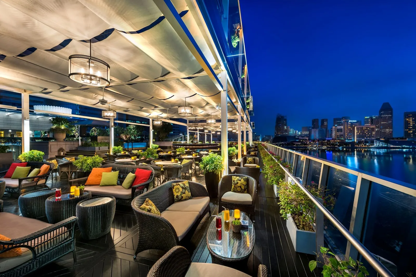 Lantern’s sofas along the glass balustrade under white awnings, with the city lit at dusk.