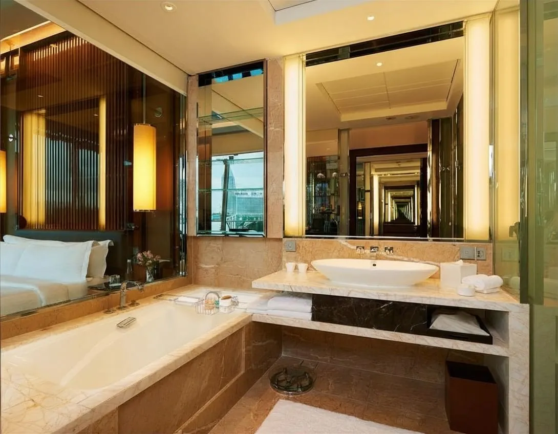 A marble bathroom: the tub below a wide mirror and the basin, with the bedroom in the reflection.