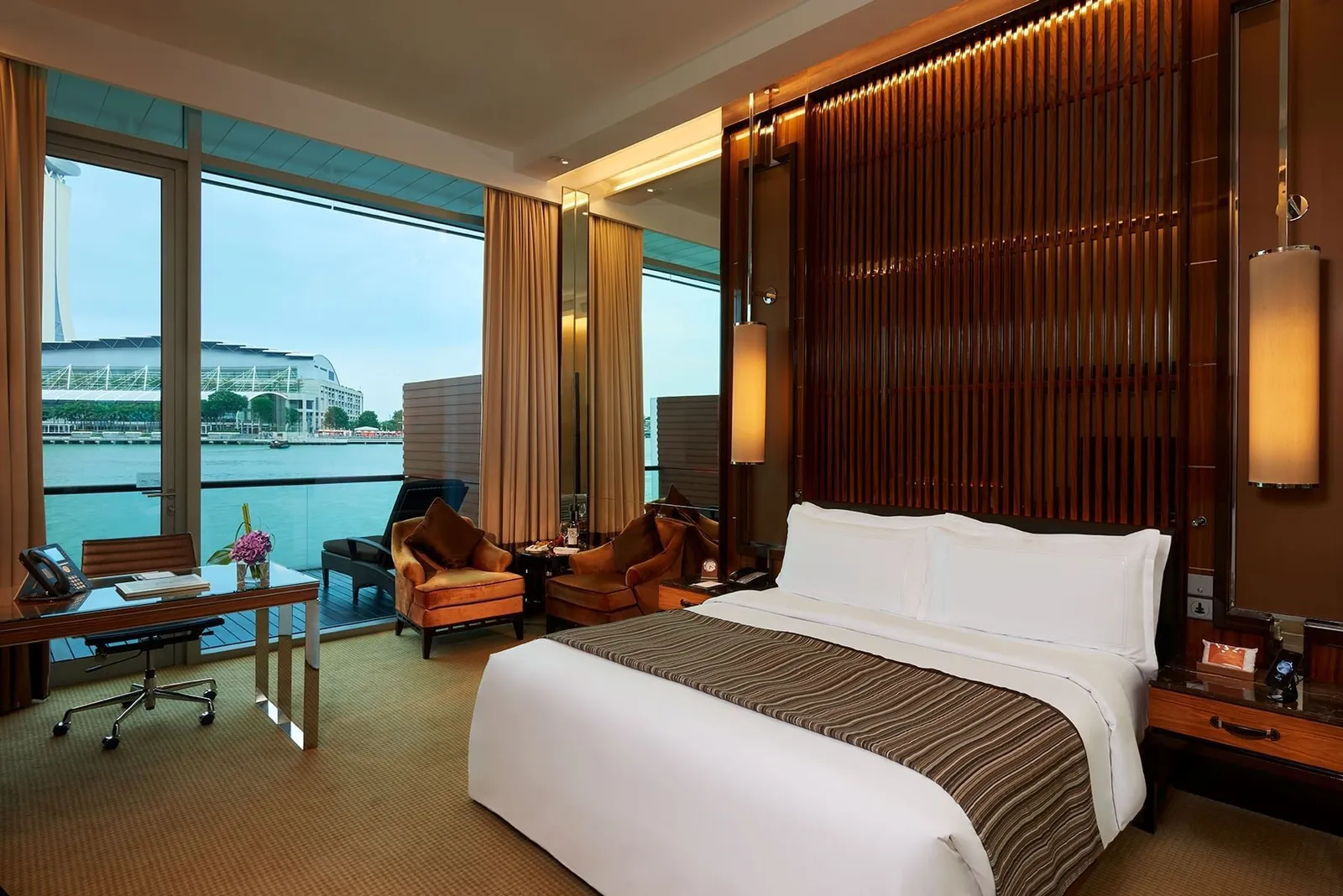 A Premier Bay View Room: the bed against a wall of dark wooden slats and armchairs by the windows onto the water.