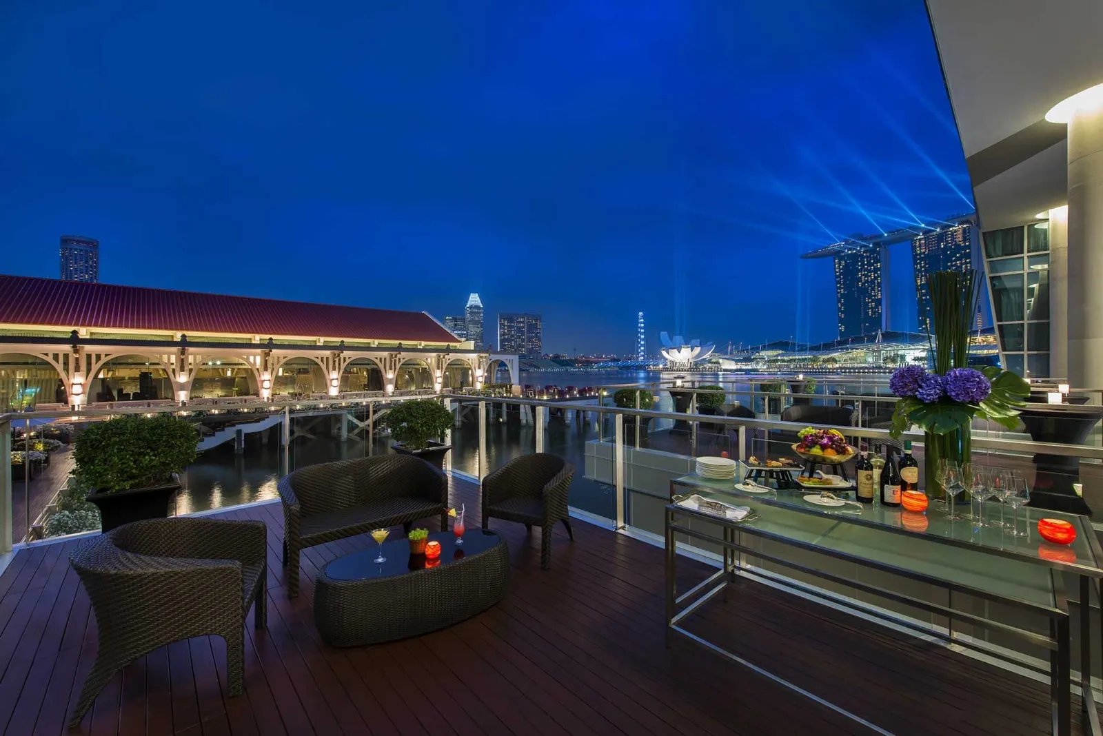 A terrace of the hotel at blue hour: Clifford Pier’s red roof, the bay and Marina Bay Sands with its beams of light.