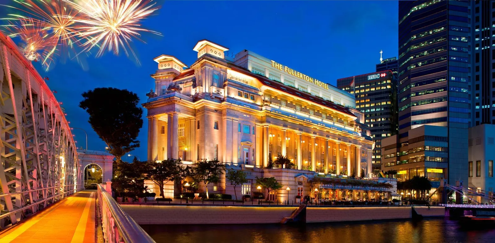 Il Fullerton Hotel illuminato al crepuscolo sulla riva del fiume, con un ponte d’acciaio a sinistra, fuochi d’artificio in cielo e i grattacieli alle spalle
