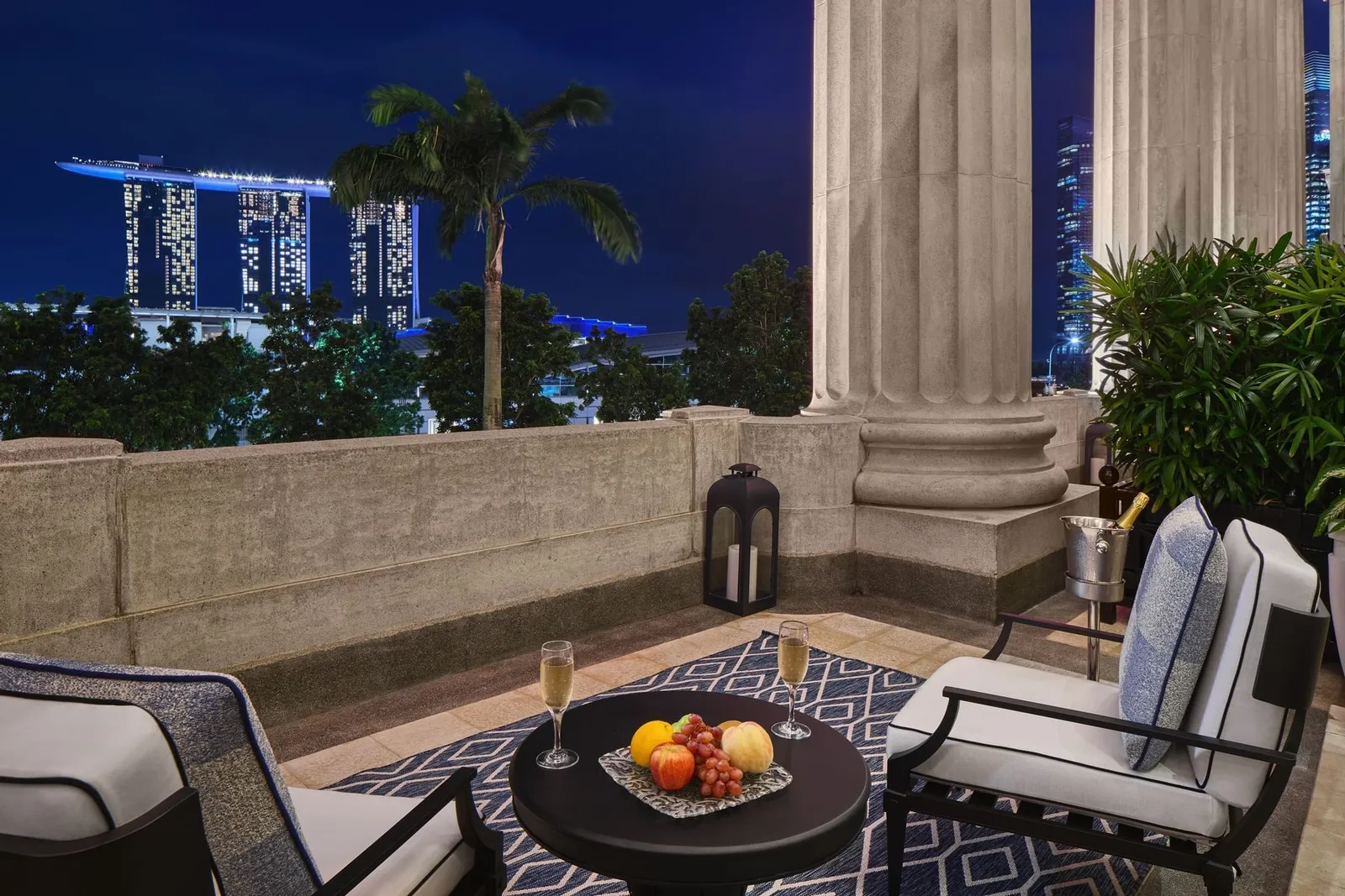 La veranda di una Loft Suite di sera: poltrone e un piatto di frutta fra le colonne doriche, con Marina Bay Sands illuminato oltre l’acqua