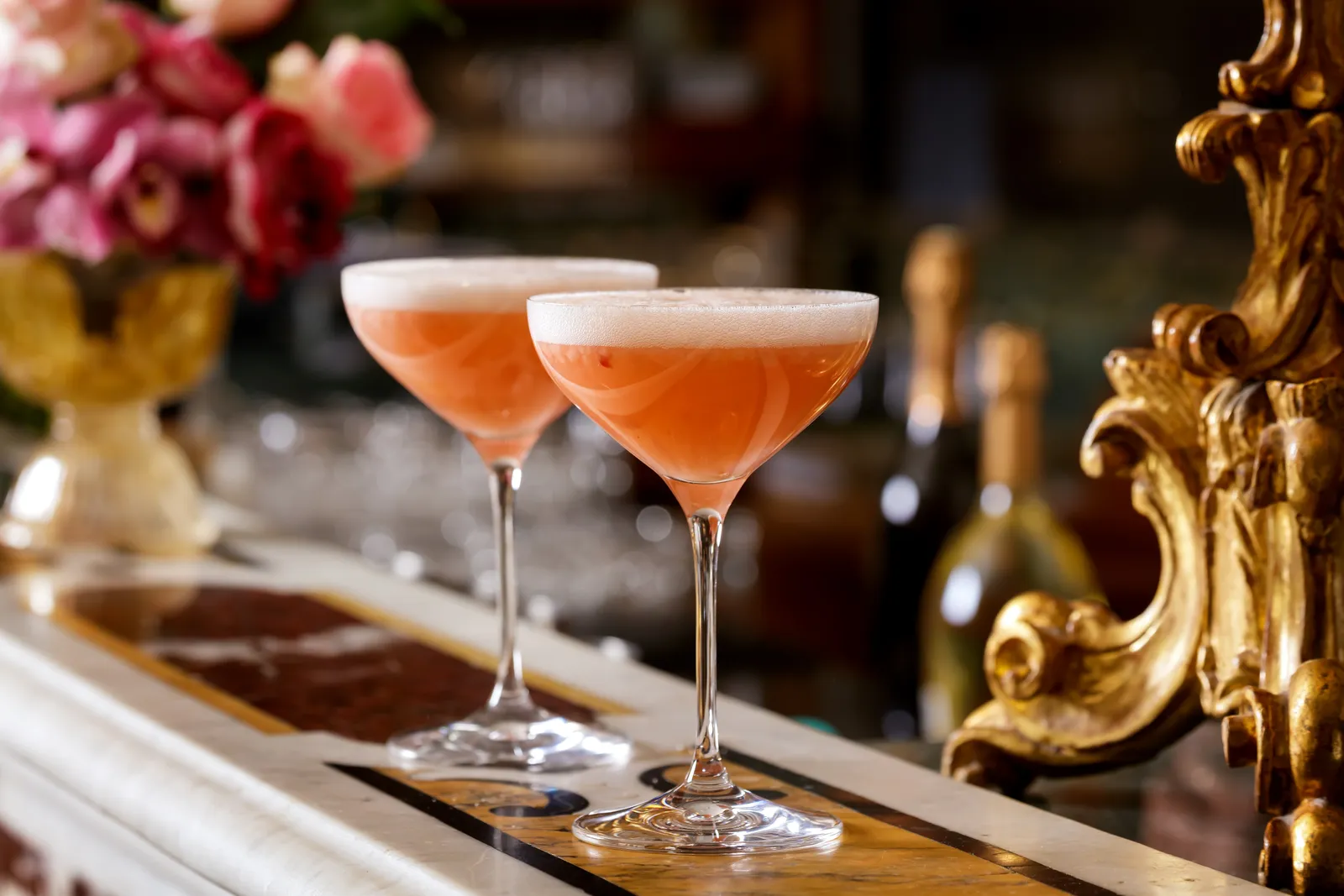 A pair of peach-coloured cocktails in stemmed coupes on the Bar Longhi counter, beside a carved gilt frame.