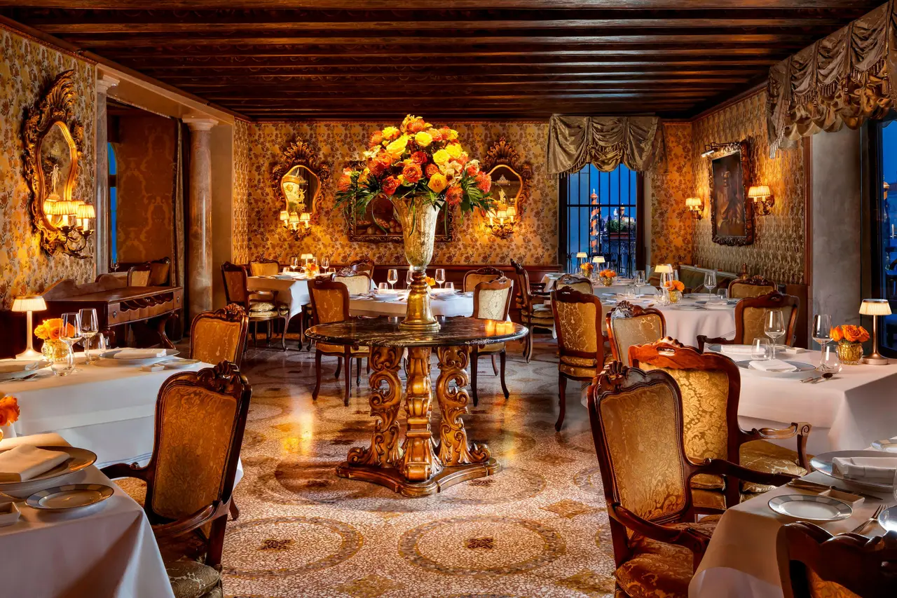 The Club del Doge dining room: white-clothed tables, gold damask walls, a tall vase of orange flowers and a beamed ceiling.