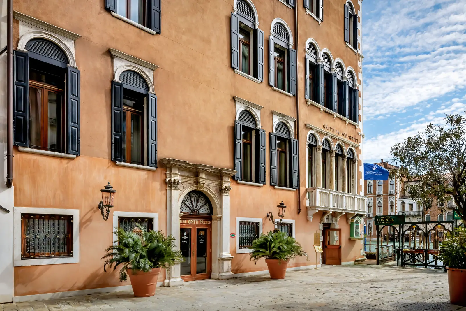 The hotel’s street entrance on the campo: the apricot façade, the stone doorway between 2 lanterns and, beyond, the Grand Canal.