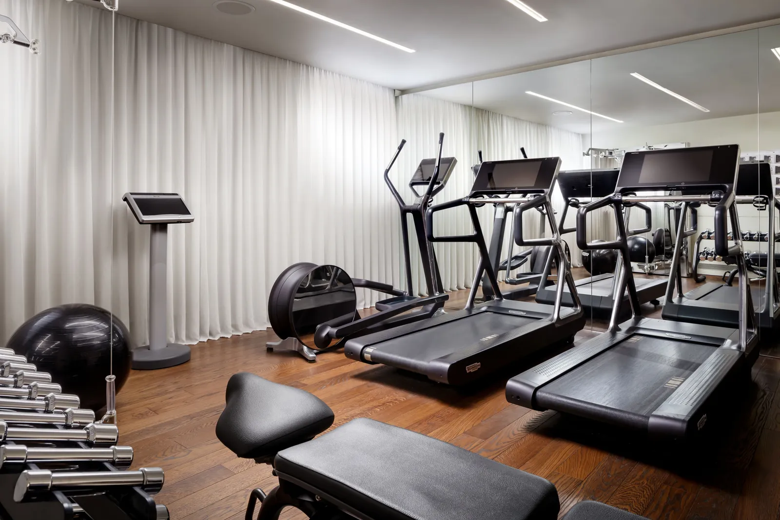 The gym: 3 treadmills and a cross-trainer before the mirrored wall, a row of dumbbells and a wooden floor.