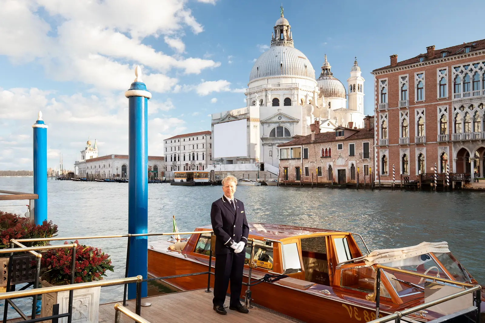 A uniformed concierge on the hotel’s landing stage beside a wooden water taxi, the Salute basilica on the far bank.
