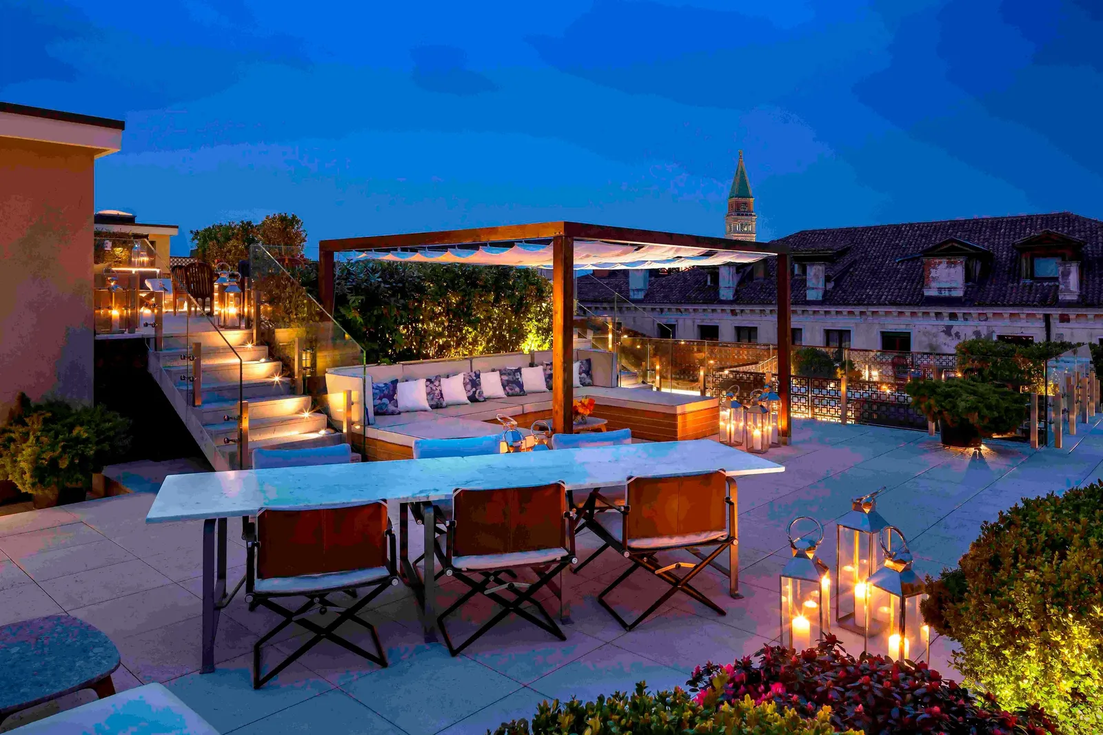 The Redentore Suite’s rooftop terrace at nightfall: a table with director’s chairs, sofas under the pergola, lit lanterns and St Mark’s bell tower.