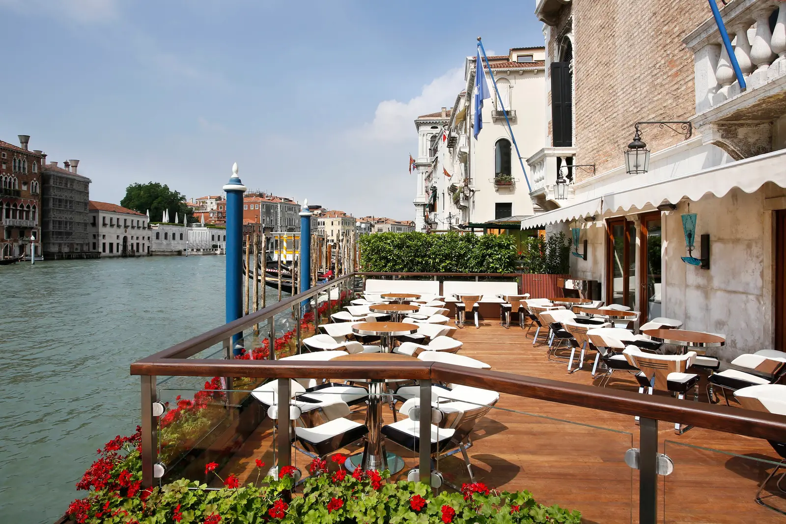 The Riva Lounge by day: white chairs and small tables on a polished wooden deck over the Grand Canal, red geraniums along the rail.