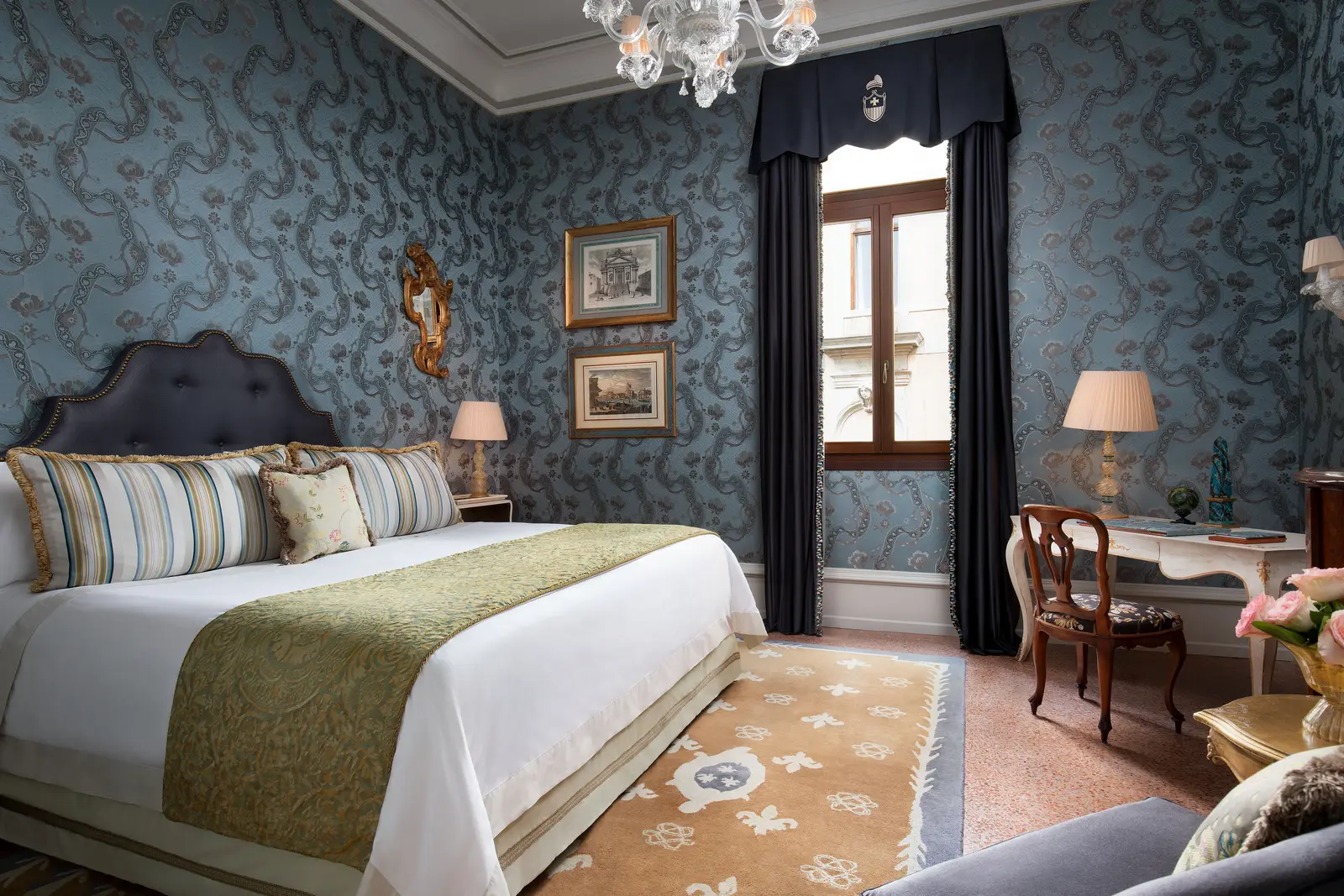 A Deluxe room: the king bed with a blue headboard, patterned pale-blue wall fabric, a writing desk and a Murano chandelier.