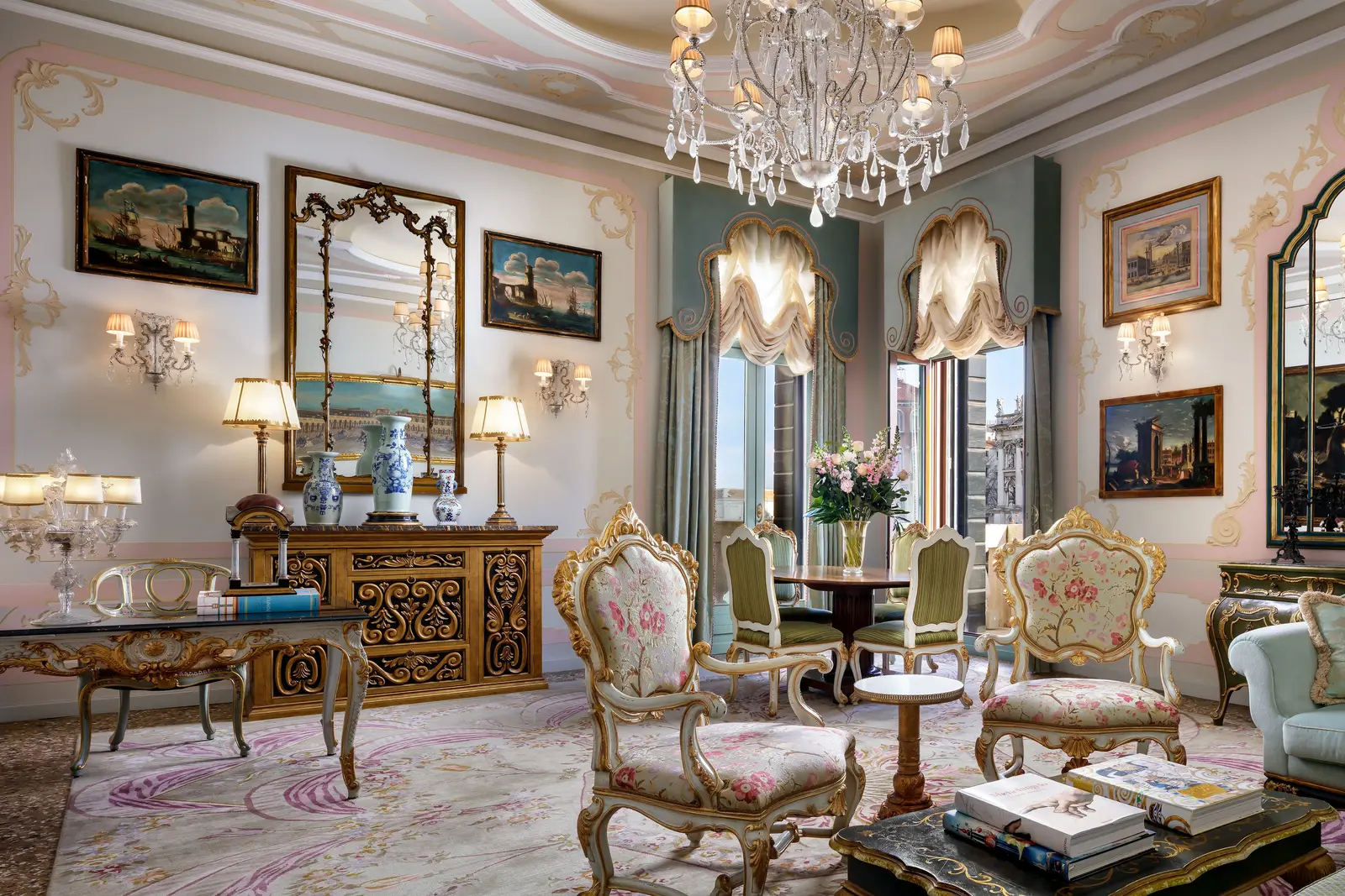 The Palazzo Canal Suite sitting room: gilt floral armchairs, Venetian mirrors, painted views and 2 windows with swagged curtains.