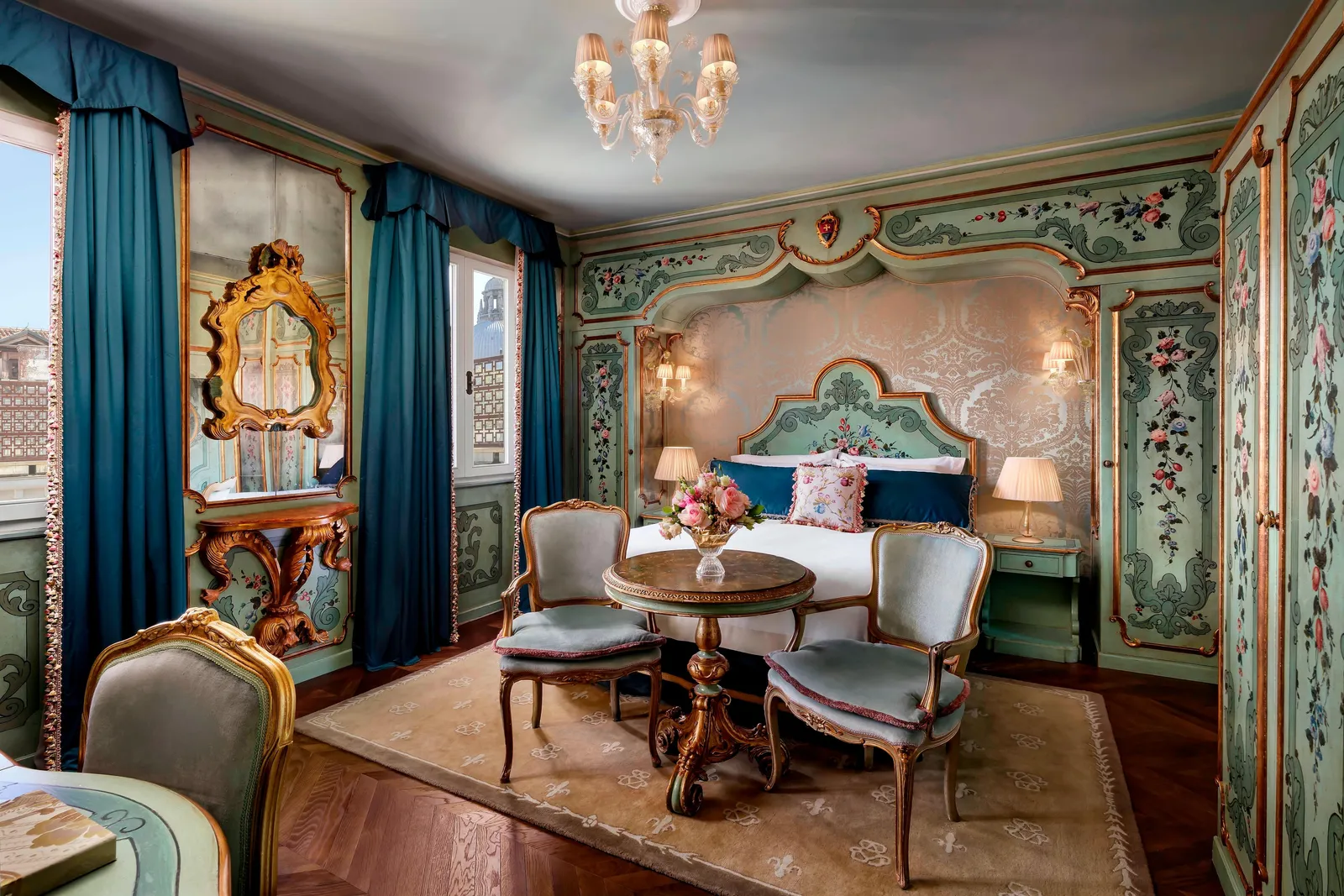 A Venetian room: the bed alcove and flower-painted wooden walls, blue curtains and a small round table with 2 chairs.