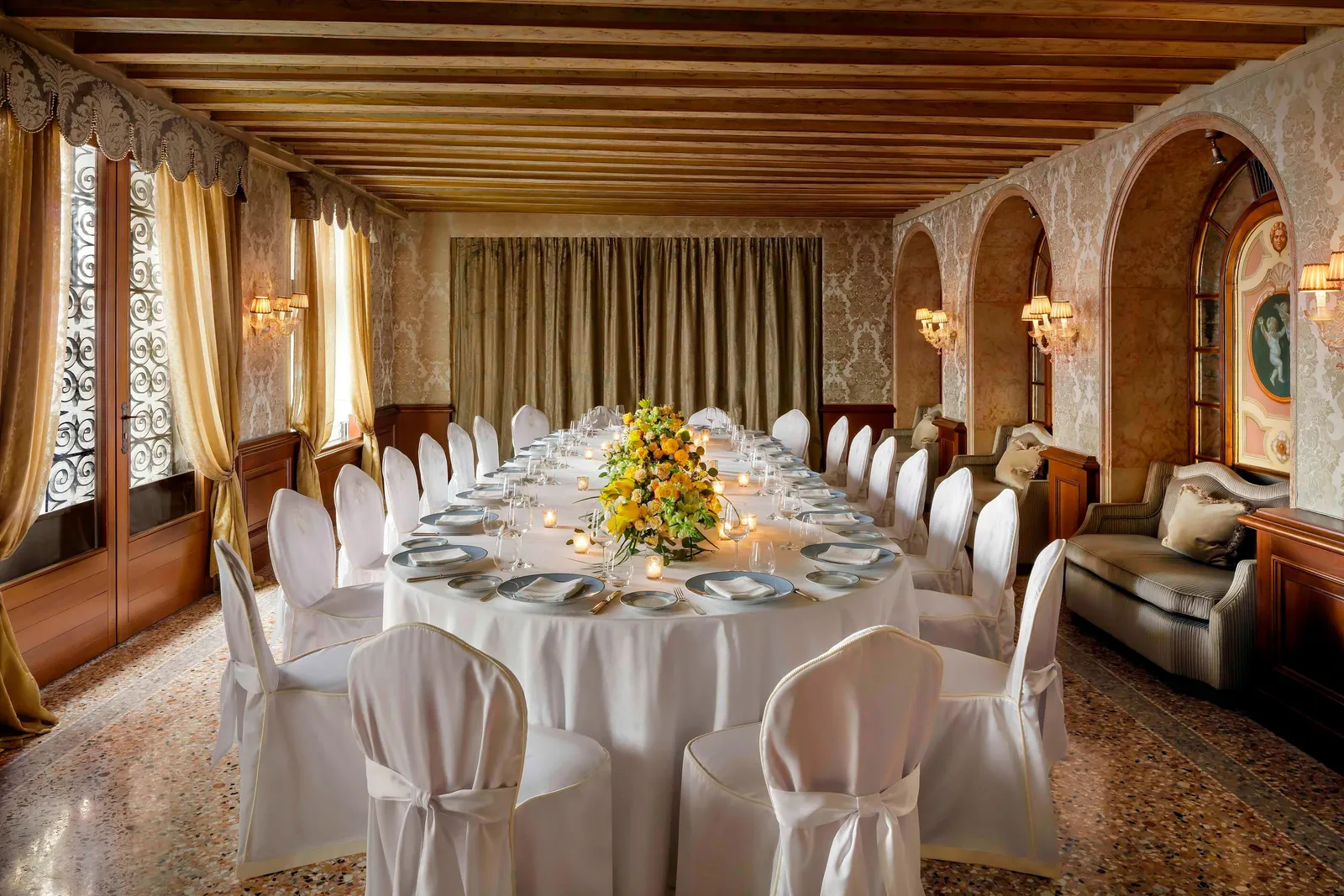 The Sala Pisani laid for a dinner: a long oval table with white-covered chairs under a beamed ceiling, between arches and damask.