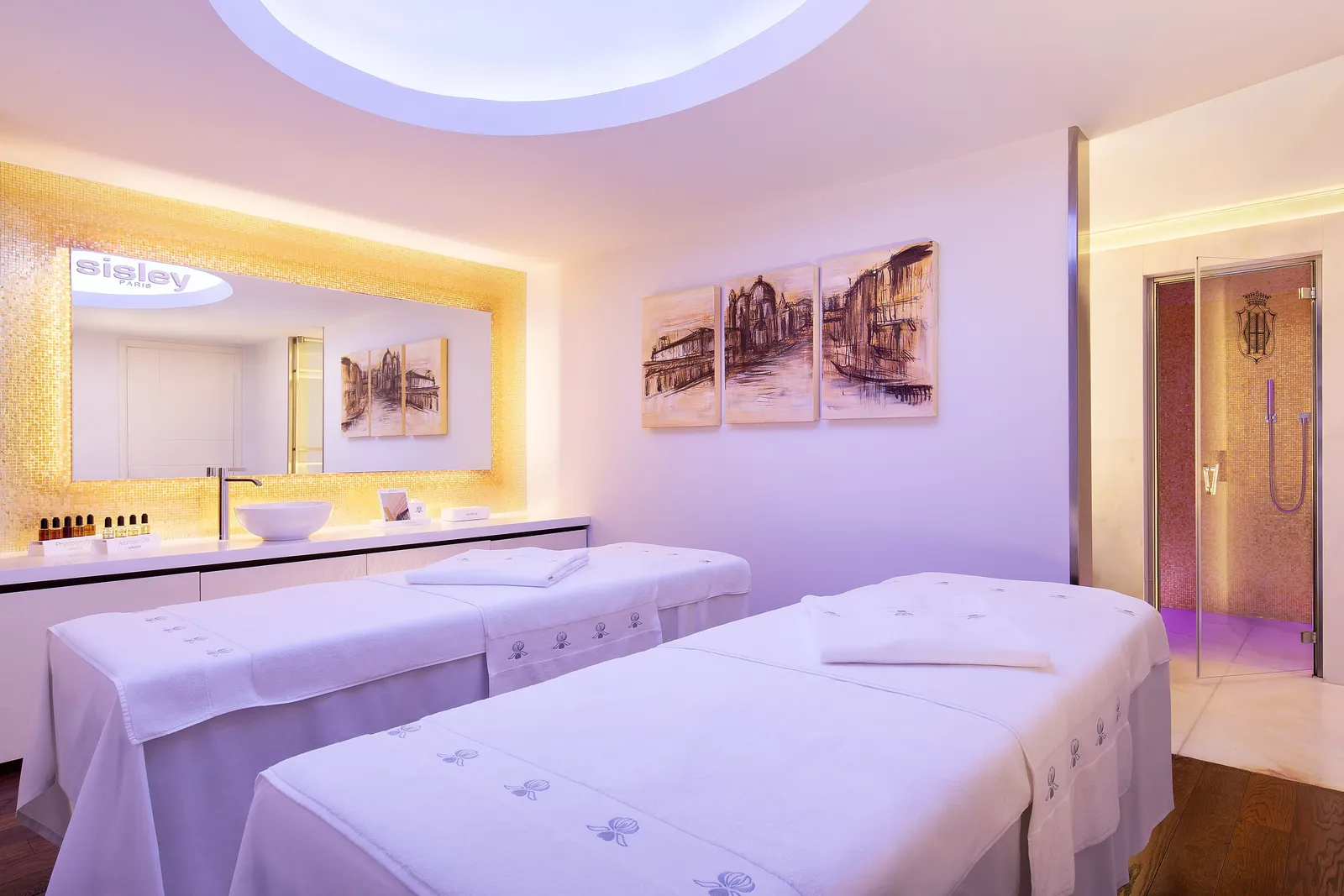 A double spa suite: 2 treatment beds under a dome of light, a gold mosaic wall and the door to the steam bath.