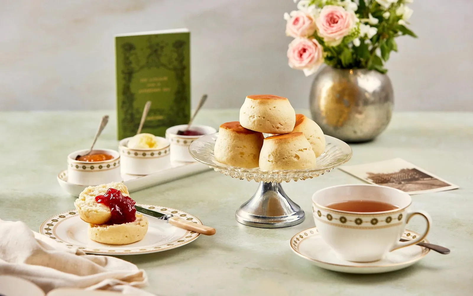 Scones with clotted cream and jam, a cup of tea and a vase of pink roses on a pale green table