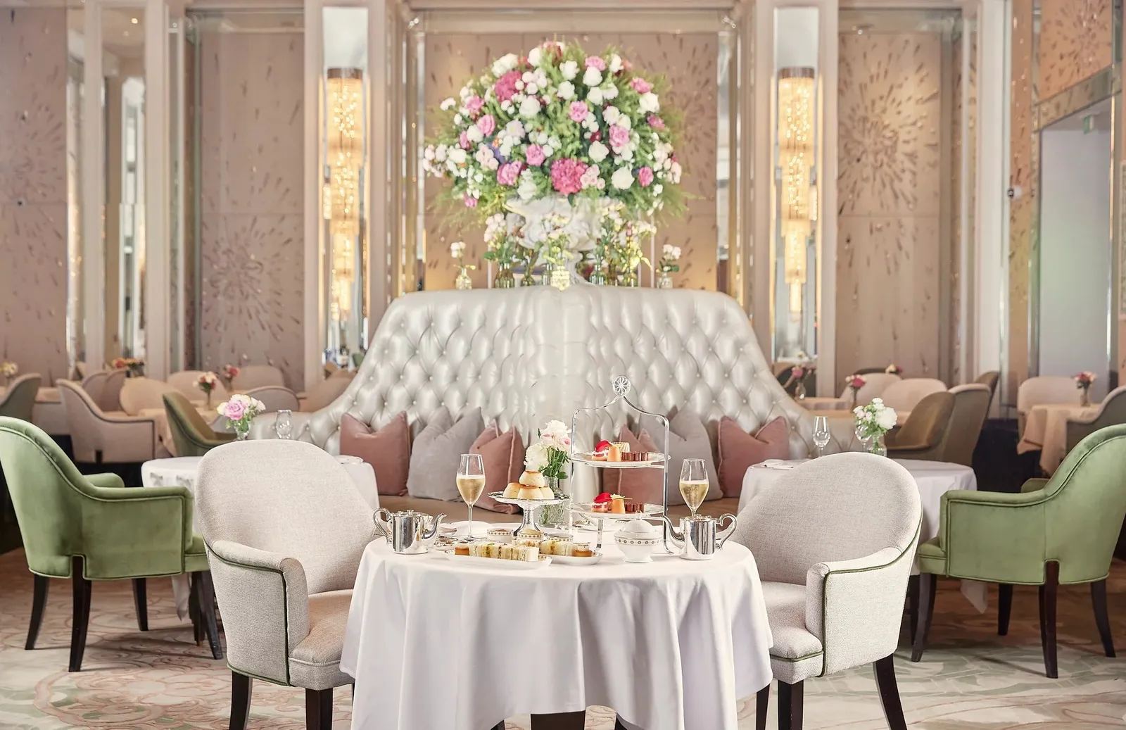 A round table laid for tea in Palm Court: a silver stand of pastries, Champagne flutes, sage-green armchairs and a tall arrangement of roses and hydrangeas