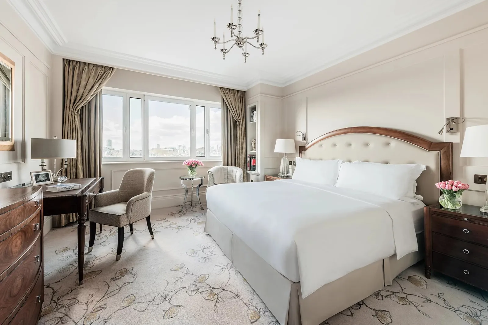 A Deluxe Room: a king bed with an upholstered headboard, a dark-wood chest, a desk and windows over the rooftops