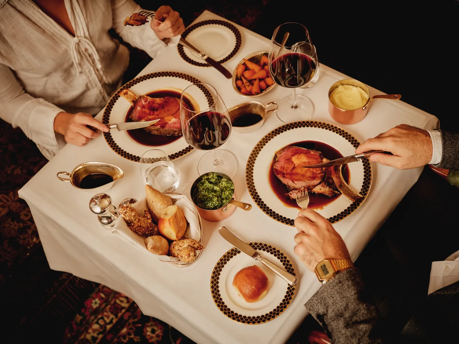 Hands carving prime rib at the table, among gold-rimmed plates and glasses of red wine