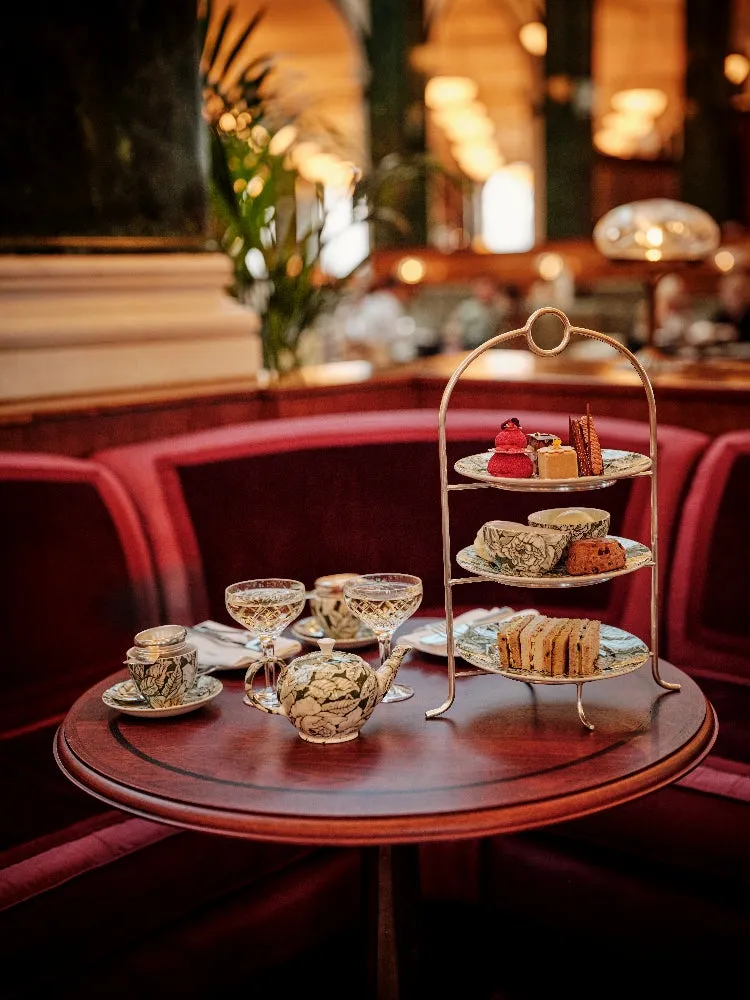 A tea stand of cakes and sandwiches on a round table, before a red velvet banquette
