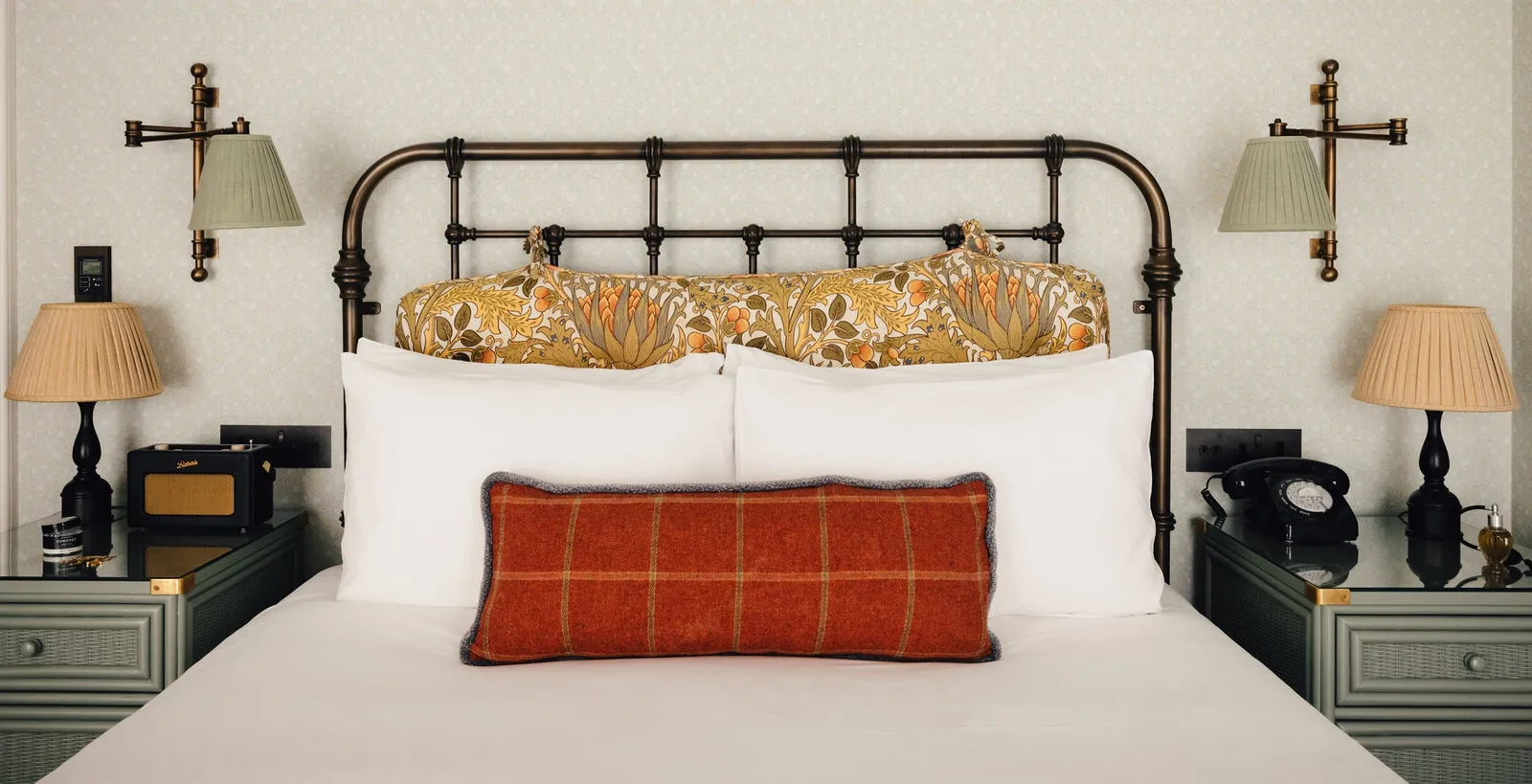 A Crash Pad: a white bed with an iron headboard, floral cushions and a red check bolster, 2 bedside lamps