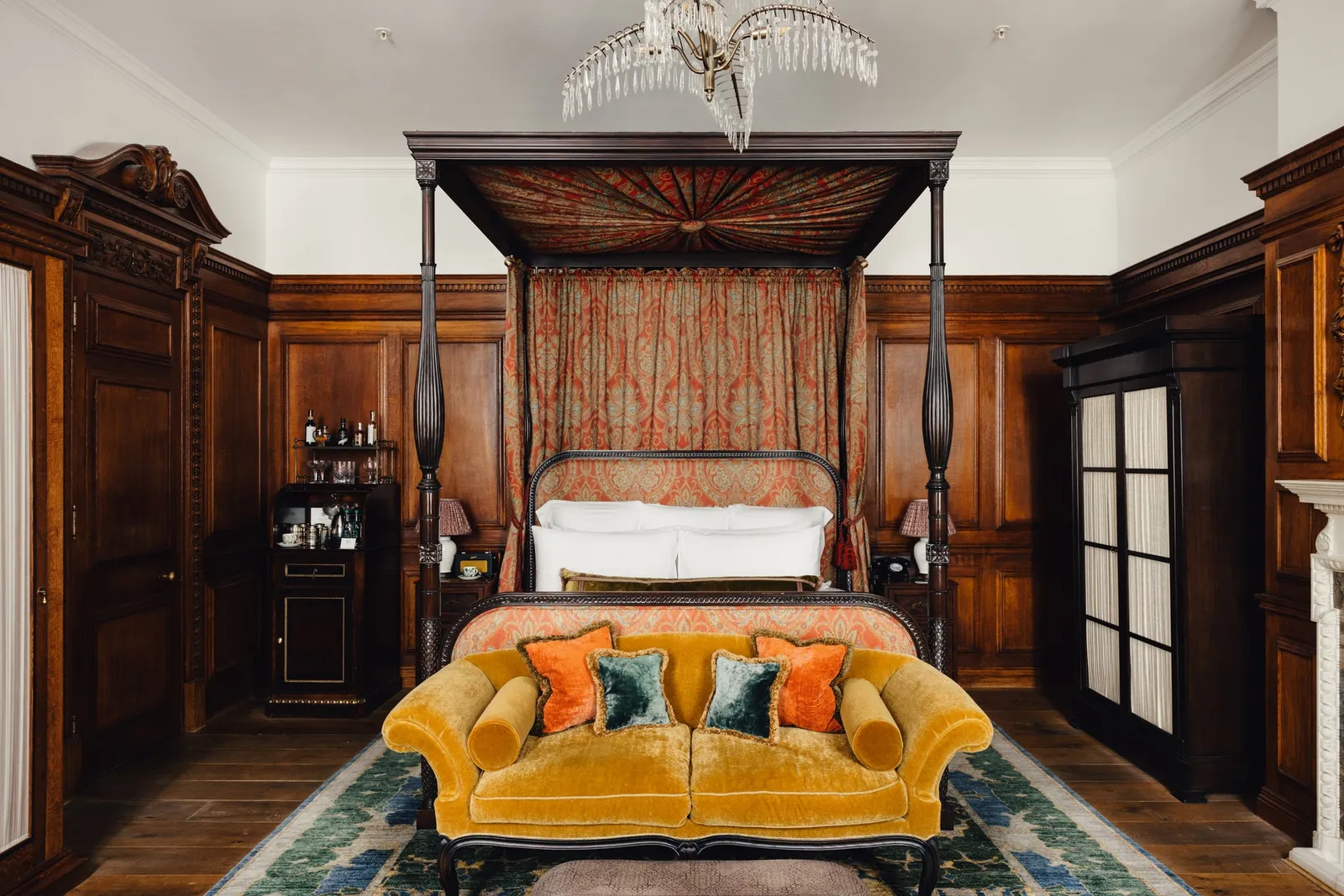 A Grand Heritage room: a canopy bed hung in red, a yellow velvet sofa, walnut panelling and a palm-leaf chandelier