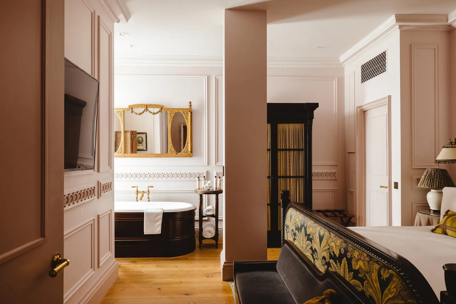 The Lutyens Suite: a pink bathroom with a roll-top bath at the end, a wooden door and a floral sofa