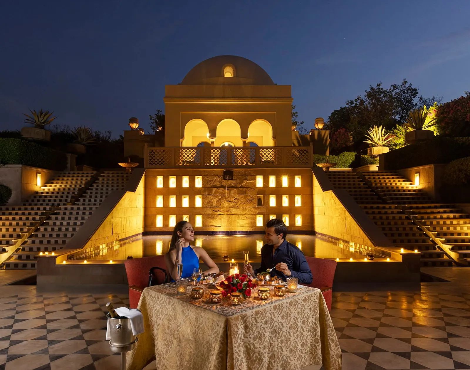 A couple dining by the pool, before the lit pavilion and the cascading water