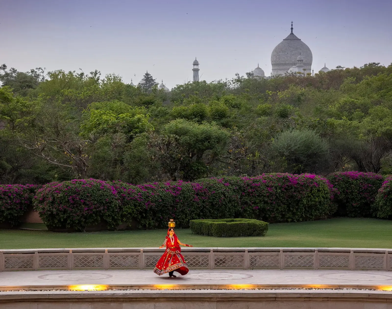 A dancer in red at the edge of the pool, with bougainvillea and the dome of the Taj Mahal behind the trees