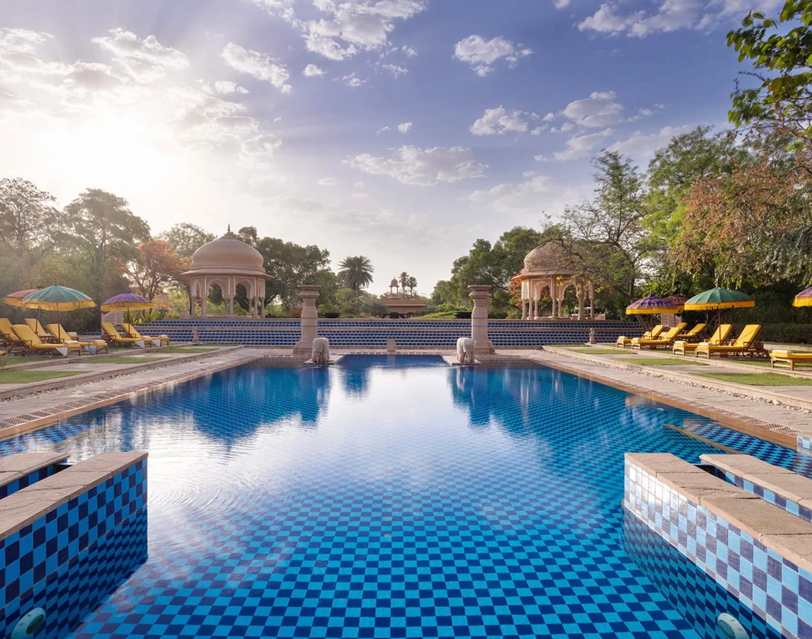 The main pool between the lawns, stone pavilions at the far end and yellow loungers along the sides