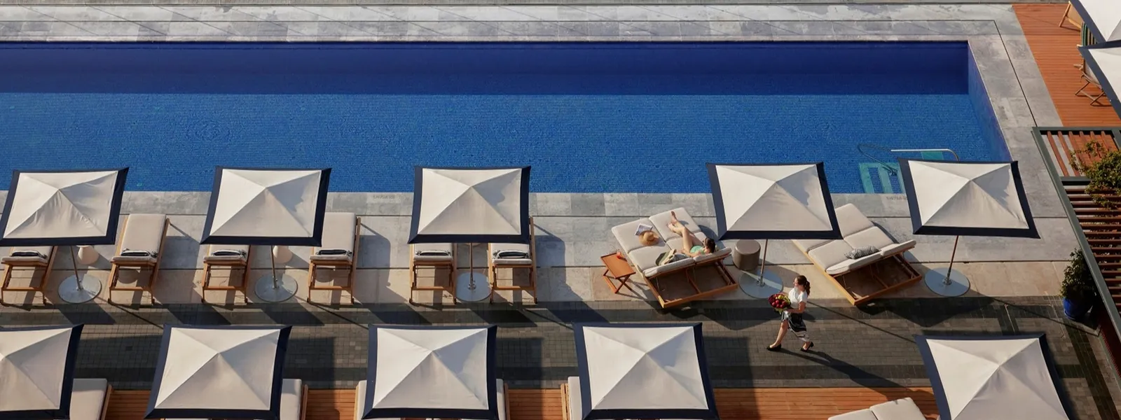 The outdoor pool from above: rows of white parasols and loungers on the deck, and a figure crossing it.